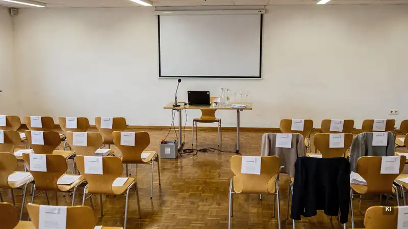 Sitzreihen mit Unterlagen für Veranstaltungsteilnehmer, Präsentationstisch mit Laptop, Mikrofon und Gläsern vor Leinwand