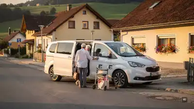 Älteres Ehepaar mit Gehhilfen steigt in weißen Kleinbus vor Wohnhäusern in ländlicher Umgebung ein.