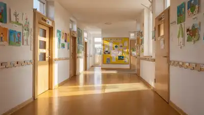 Heller Schulflur mit Holztüren, Kinderzeichnungen an den Wänden, Garderobenhaken und gelber Infotafel am Ende des Gangs.