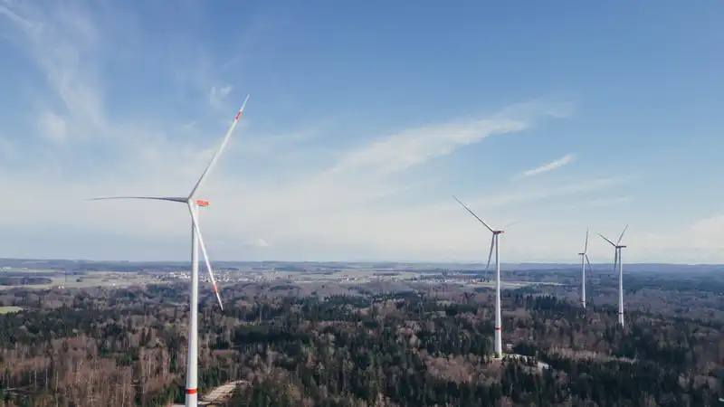 Windkraftanlagen auf bewaldeten Hügeln unter blauem Himmel mit weißen Wolken in ländlicher Landschaft